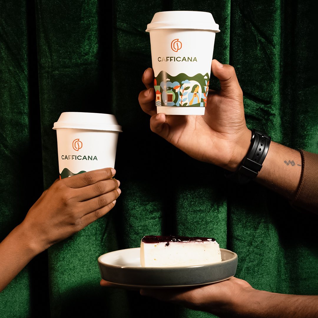 Two Cafficana paper cups with illustrated farm landscape, held against deep green velvet